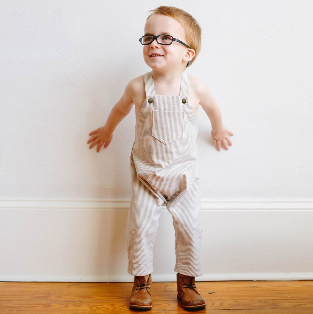 Photo of 2 year old red head boy wearing Beya Made engineer stripe overalls standing against a white wall