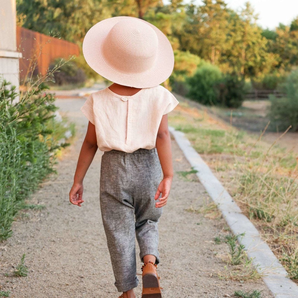 back view of Beya Made linen top worn by 3 year old girl with sun hat, walking along a meadow path