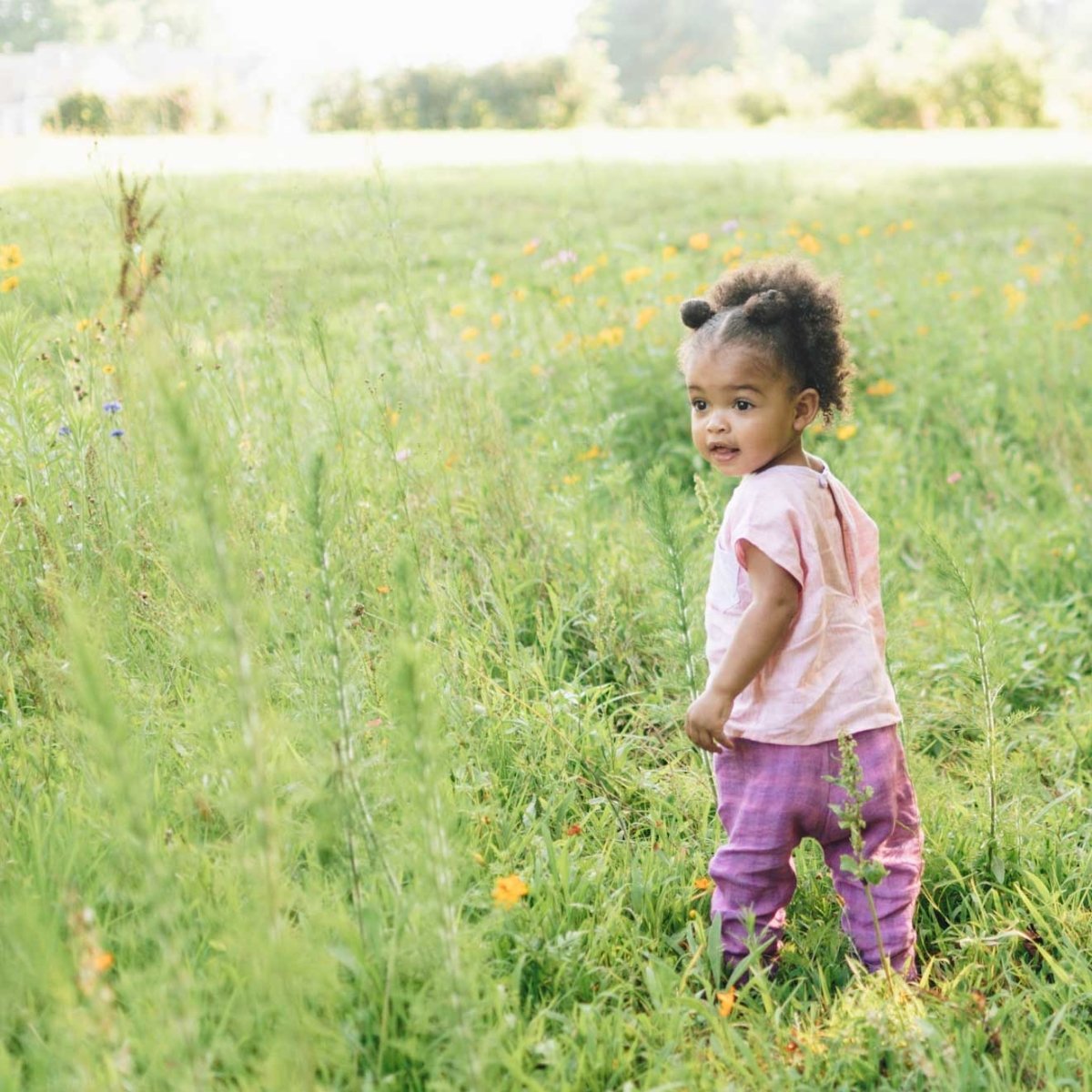 2 year old girl wearing pink Beya Made top and purple pants, standing in a meadow
