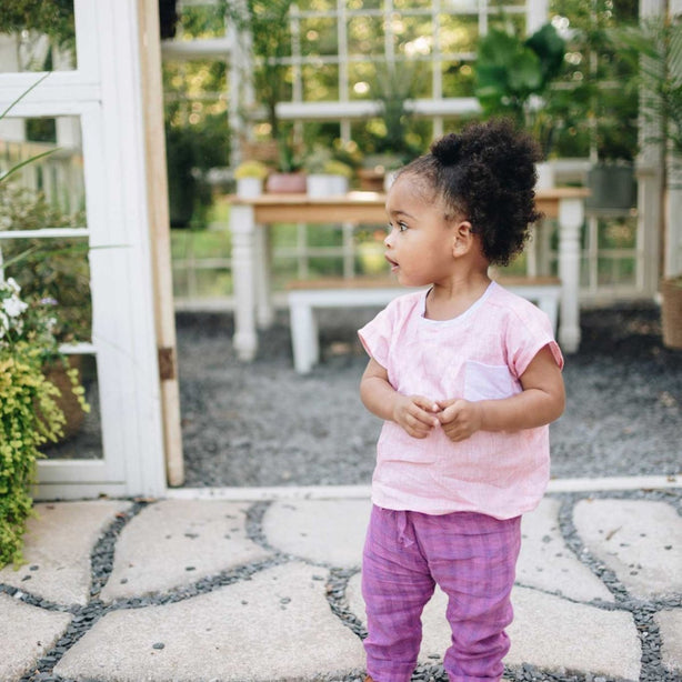 Little girl wearing pink linen top and purple linen pants standing in front of greenhouse