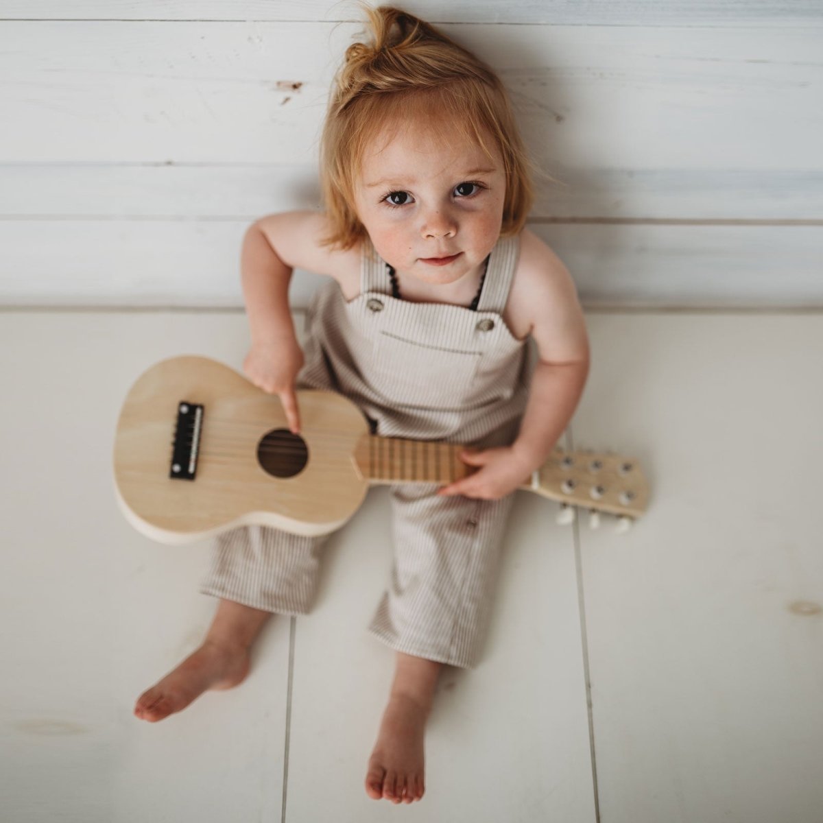 Photo of toddler boy wearing a beige linen romper sitting on a wood floor playing a ukelele
